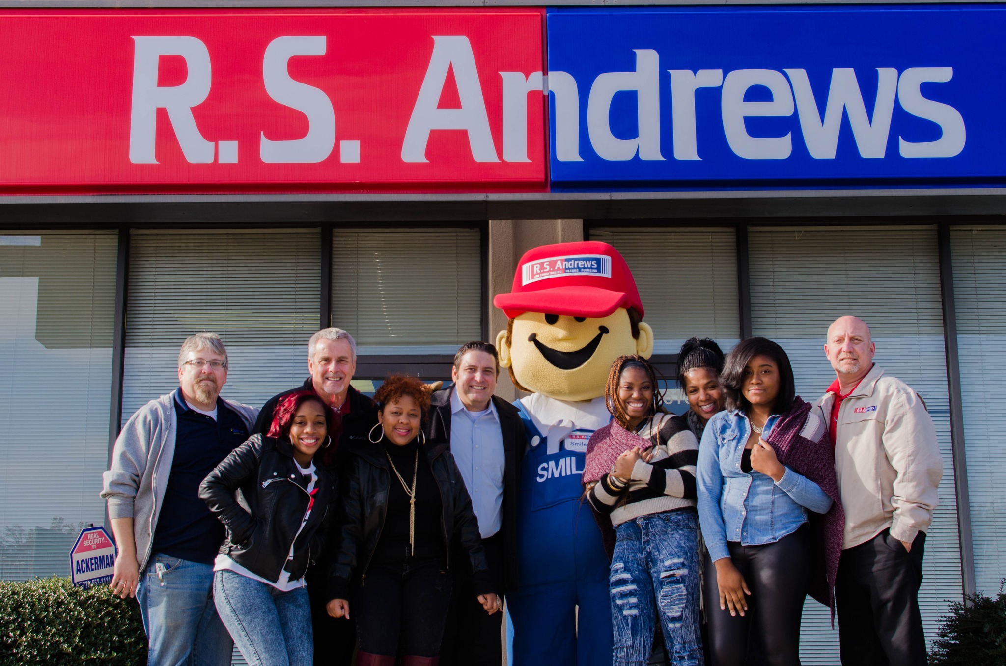 Group of diverse coworkers posing with a smiling mascot in front of a red-and-blue RS Andrews sign building entrance, outdoors, daytime.